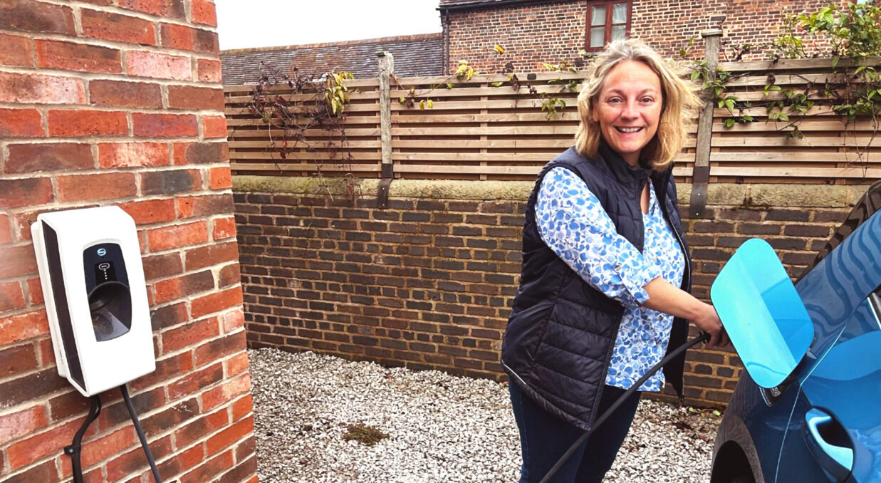 A woman in a blue patterned shirt and gilet is pluggin a blue EV into an Indra Smart PRO charger