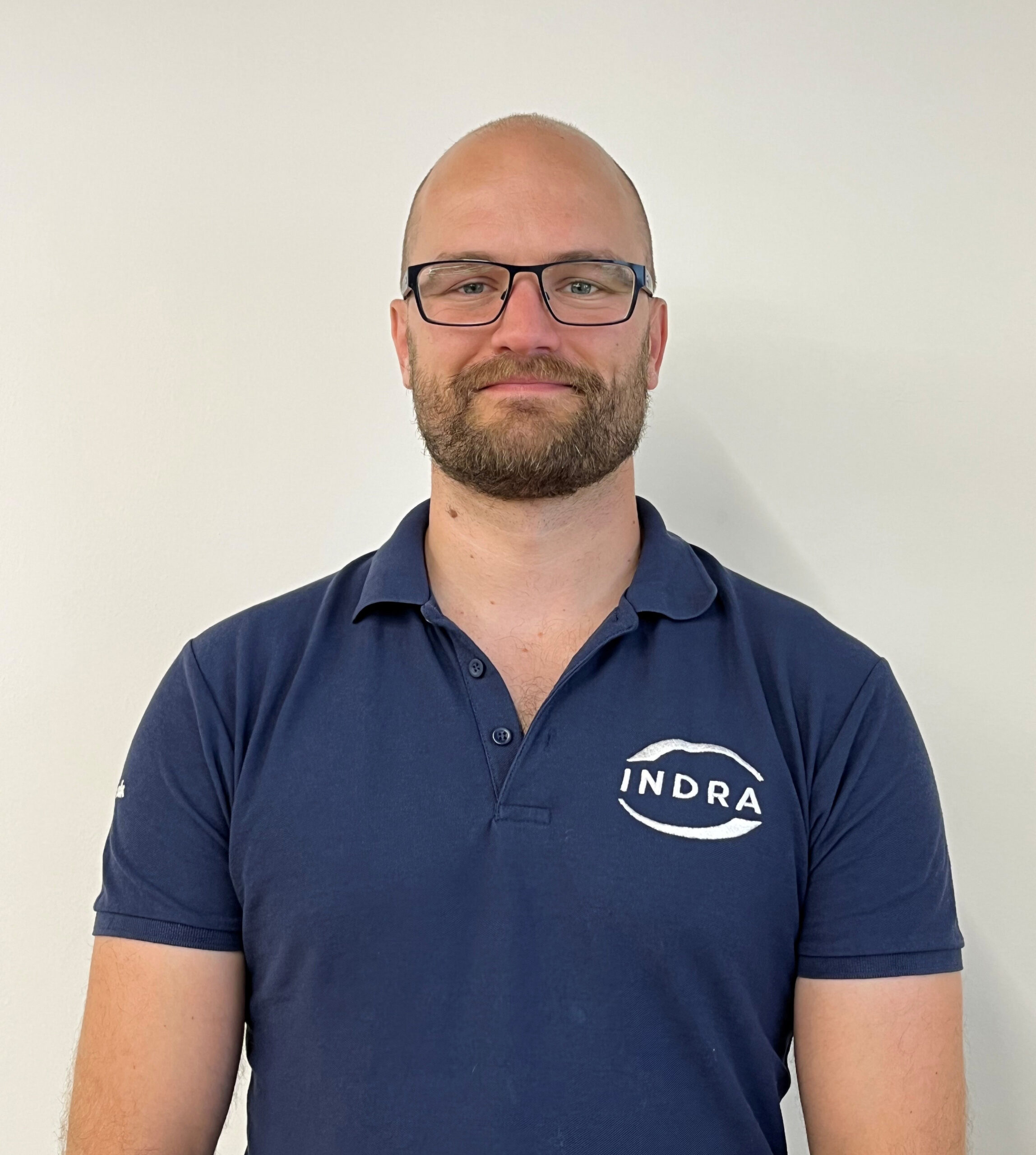 Man with beard and glasses and blue Indra logo polo shirt smiles at camera